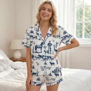 Puede incluir: Conjunto de pijama blanco con estampado azul de tem&aacute;tica occidental. La parte superior de manga corta y los pantalones cortos presentan ilustraciones de caballos, vaqueros, cactus y ganado. El conjunto tiene ribetes azules y cierre de botones.