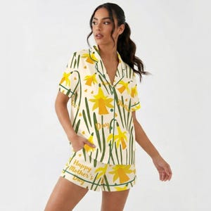 May include: Cream-colored pajama set with a daffodil pattern and green trim. The short-sleeved top and shorts feature yellow daffodils and green stems. Text on the shorts reads "Happy Mother's Day".