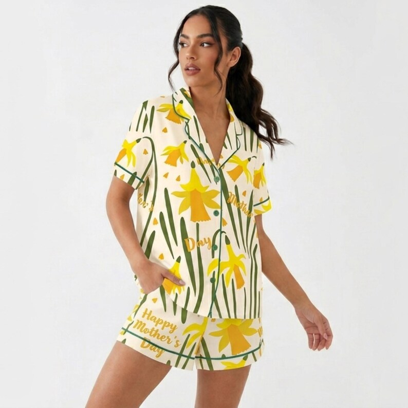May include: Cream-colored pajama set with a daffodil pattern and green trim. The short-sleeved top and shorts feature yellow daffodils and green stems. Text on the shorts reads "Happy Mother's Day".