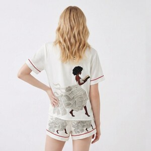 May include: White pajama set with red trim, featuring a graphic of a woman reading, her dress made of newspaper clippings. The short-sleeved top and shorts create a coordinated sleepwear ensemble.