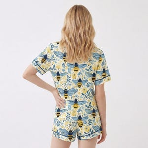 May include: A two-piece pajama set featuring a repeating pattern of bees and floral elements on a cream background. The short-sleeved top and shorts have navy blue trim. The bee design includes yellow and black stripes with blue wings.