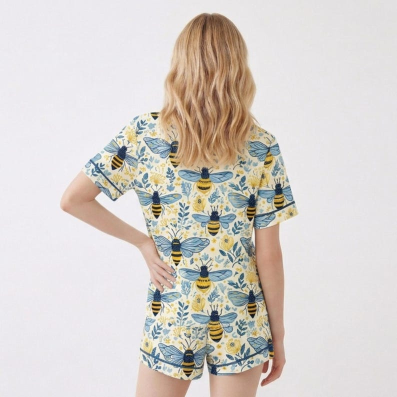 May include: A two-piece pajama set featuring a repeating pattern of bees and floral elements on a cream background. The short-sleeved top and shorts have navy blue trim. The bee design includes yellow and black stripes with blue wings.