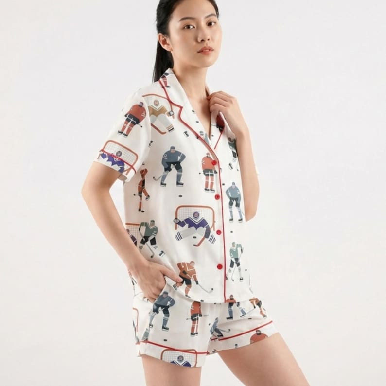 K&ouml;nnte beinhalten: Wei&szlig;es Pyjama-Set mit Hockey-Motiv. Das kurz&auml;rmelige Oberteil und die Shorts zeigen ein Muster aus Hockeyspielern, Netzen und Pucks in verschiedenen Farben. Rote Paspeln s&auml;umen die Kanten des Sets und setzen einen Kontrast.