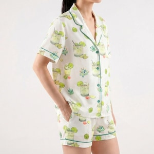 May include: White pajama set with a green trim, featuring a repeating pattern of cocktails, limes, and cacti. The short-sleeved top and shorts are made of a lightweight fabric. The set is designed for comfort and relaxation.