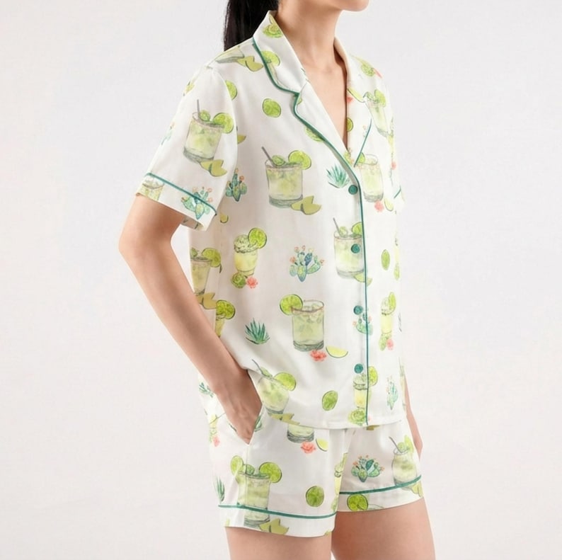 May include: White pajama set with a green trim, featuring a repeating pattern of cocktails, limes, and cacti. The short-sleeved top and shorts are made of a lightweight fabric. The set is designed for comfort and relaxation.