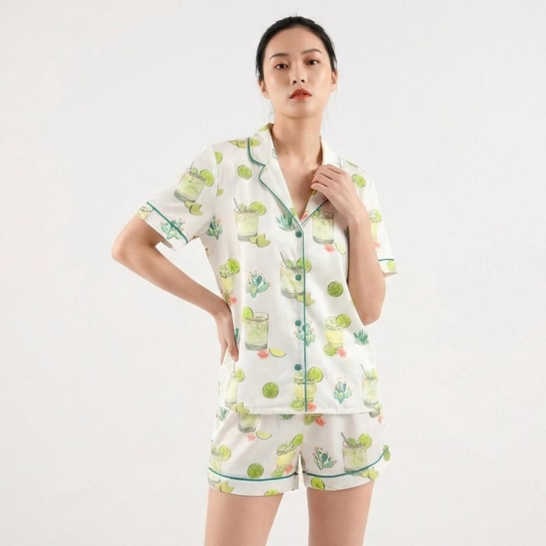 May include: White pajama set with a green and lime cocktail print. The short-sleeved top and shorts have green trim. The fabric features illustrations of cocktails, limes, and sprigs of greenery.