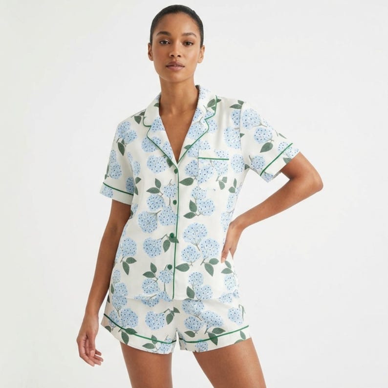 May include: A matching pajama set featuring a short-sleeved button-down shirt and shorts. The set is white with a blue floral pattern and green trim. The model is wearing the pajama set.