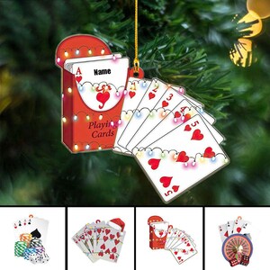 May include: A red and white playing card ornament with a deck of cards and a fan of cards. The deck of cards has the word "Name" on it. The cards are decorated with hearts and lights. The words "Playing Cards" are on the box.