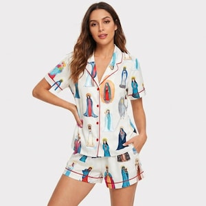 Könnte beinhalten: Weißes Pyjama-Set mit einem Muster religiöser Figuren in verschiedenen Farben. Das kurzärmelige Oberteil und die Shorts haben rote Paspeln und Knöpfe. Das Pyjama-Set ist aus einem weichen, bequemen Material gefertigt.