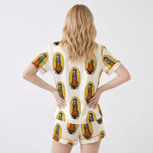 Pode incluir: Conjunto de pijama creme com um padr&atilde;o repetido de uma figura religiosa em uma moldura oval. A blusa de manga curta e os shorts t&ecirc;m acabamento laranja. A figura &eacute; retratada em azul, laranja e amarelo.