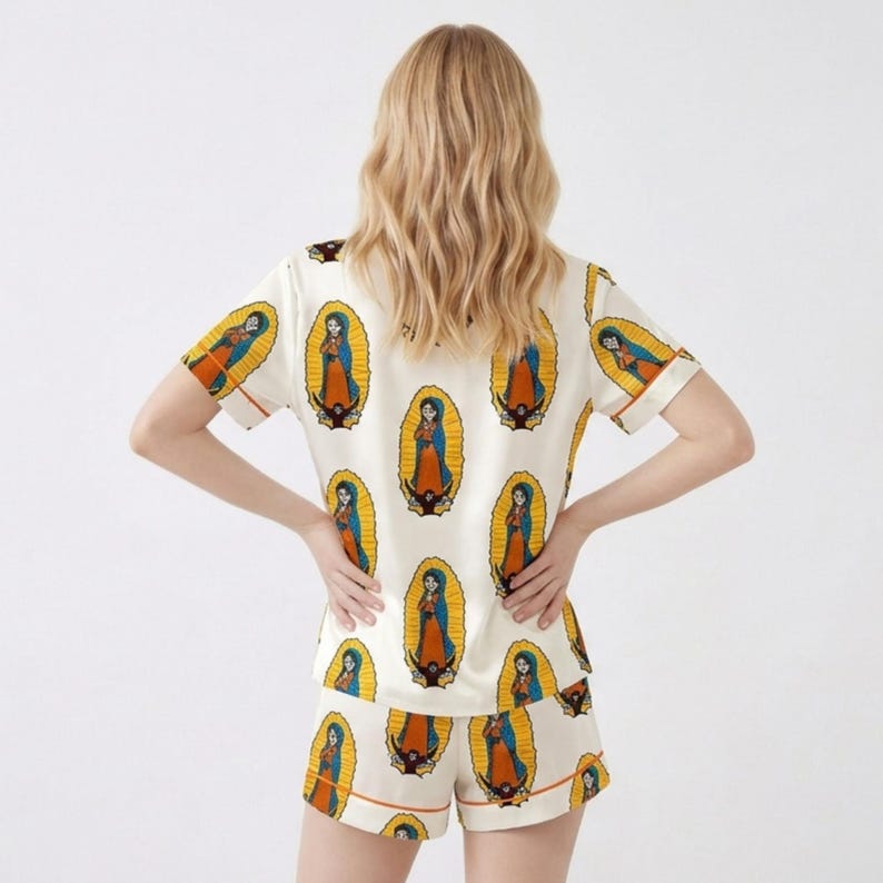 Pode incluir: Conjunto de pijama creme com um padr&atilde;o repetido de uma figura religiosa em uma moldura oval. A blusa de manga curta e os shorts t&ecirc;m acabamento laranja. A figura &eacute; retratada em azul, laranja e amarelo.