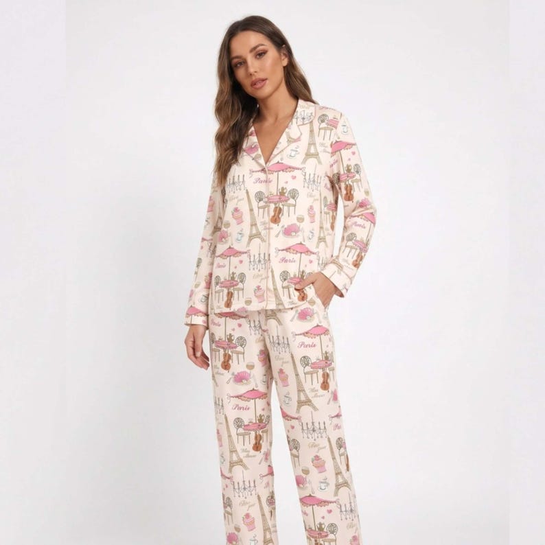 May include: A light pink two-piece pyjama set with a repeating pattern of Parisian cafe scenes, including the Eiffel Tower, tables, and umbrellas. The long-sleeved top has a button-down front and a notched collar. The trousers are straight-legged.