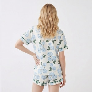 May include: Cream-colored pajama set with a blue floral pattern. The short-sleeved top and shorts have green trim. The floral design features clusters of blue flowers and green leaves.