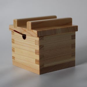 May include: A small, light-coloured wooden box with a lid and two parallel handles. The box is constructed with interlocking joints and has a small hole on the side. The box is likely used for storage or as a container.