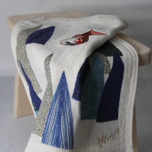 May include: A folded white towel with an abstract design in shades of blue, green, and gray. The towel features a stylized illustration of a figure and a signature in gold thread. The towel is draped over a light wooden stool.