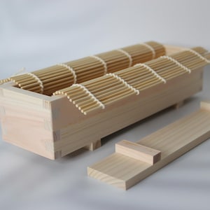 May include: A wooden sushi making kit with a rectangular box, bamboo mats, and a separate wooden piece. The box has a rectangular shape with bamboo slats on top. The kit is made of light-colored wood.