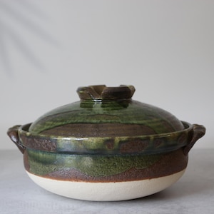 Może przedstawiać: Ceramiczny garnek do gotowania z pokrywką. Garnek jest mieszanką zieleni, brązu i bieli, z teksturowanym wykończeniem. Ma dwa uchwyty i pokrywkę z unikalnym uchwytem. Garnek jest przeznaczony do gotowania.