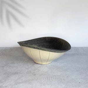Puede incluir: Un cuenco de cerámica en forma de lágrima con un exterior de color crema moteado y finas líneas verticales negras. El interior del cuenco tiene un acabado de esmalte gris carbón oscuro y texturizado, creando un contraste con la base clara.