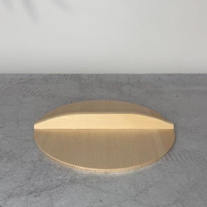 May include: A circular light-colored wooden lid featuring a straight, raised handle across the center. The wood grain is visible throughout the surface, showcasing a natural, smooth finish on the flat, round disc and the rectangular handle piece.