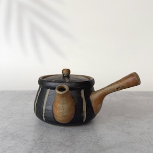 May include: A ceramic teapot with a dark black body featuring vertical light-coloured stripes. The teapot has a side handle and a spout, both finished in a natural tan clay colour. A matching circular lid sits on top of the main vessel.
