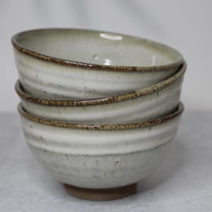 Puede incluir: Una pila de tres cuencos de cerámica hechos a mano. Los cuencos son de color gris claro con un diseño texturizado y acanalado. Los bordes de los cuencos tienen un borde marrón oscuro y rústico. Los cuencos están apilados.