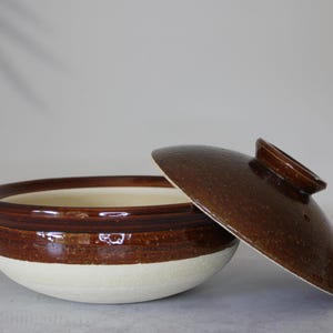 Peut inclure: Un pot de cuisson en c&eacute;ramique avec un couvercle, pr&eacute;sentant un design bicolore. Le pot a une base blanche et une section sup&eacute;rieure marron, avec un couvercle marron assorti. Le couvercle est l&eacute;g&egrave;rement entrouvert.