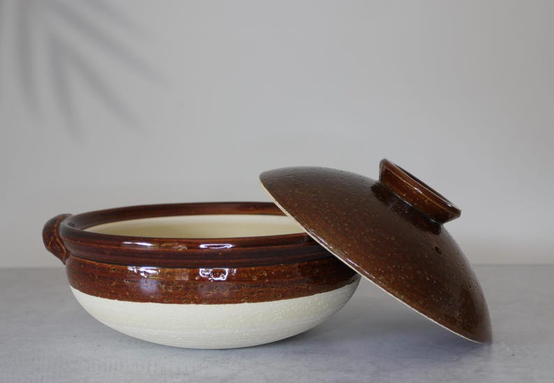 Peut inclure: Un pot de cuisson en c&eacute;ramique avec un couvercle, pr&eacute;sentant un design bicolore. Le pot a une base blanche et une section sup&eacute;rieure marron, avec un couvercle marron assorti. Le couvercle est l&eacute;g&egrave;rement entrouvert.