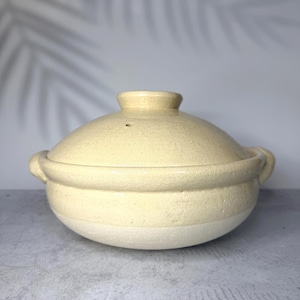 May include: A round, cream-coloured ceramic pot with a matching lid and two side handles. The pot features a textured, glazed finish on the upper portion and a matte, unglazed base. A small steam vent is visible on the top of the domed lid.