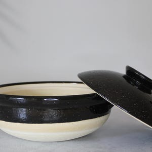 Peut inclure: Un pot de cuisson en c&eacute;ramique noir et cr&egrave;me avec un couvercle assorti. Le pot a une forme arrondie avec un bord noir et une base cr&egrave;me. Le couvercle est noir avec une finition mouchet&eacute;e. Le pot a deux petites poign&eacute;es.