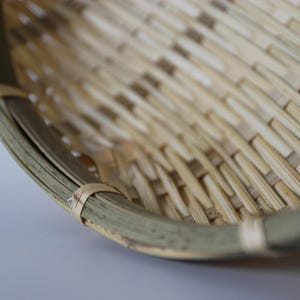 May include: A round, shallow basket constructed from woven light-colored bamboo strips. The rim is reinforced with a thicker, curved bamboo band secured by thin, light-colored bamboo ties spaced evenly along the edge of the basket structure.