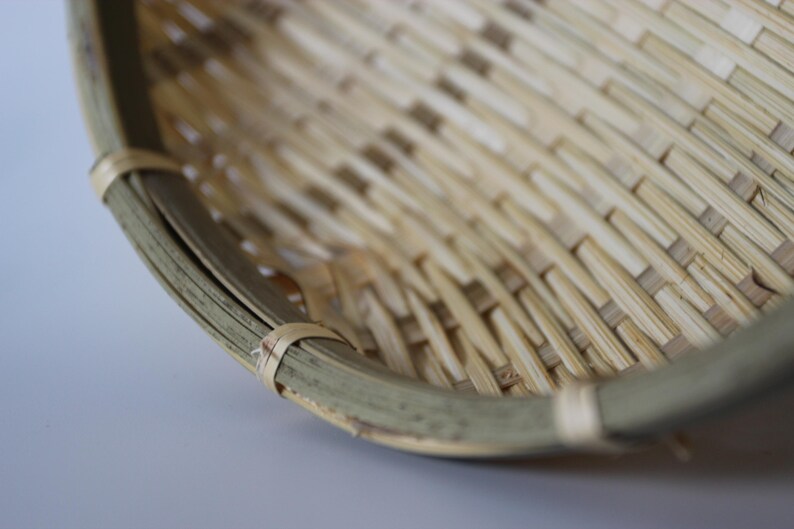 May include: A round, shallow basket constructed from woven light-colored bamboo strips. The rim is reinforced with a thicker, curved bamboo band secured by thin, light-colored bamboo ties spaced evenly along the edge of the basket structure.
