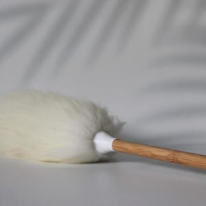 Peut inclure: Un plumeau doté d'une tête duveteuse, blanc cassé et à la texture douce, fixée à un manche en bois brun clair lisse. Le point de jonction entre la tête et le manche est une virole en plastique blanc simple qui maintient les fibres en place.