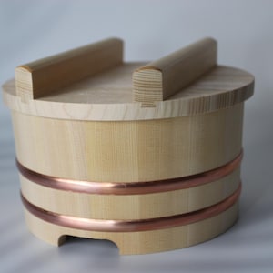 Könnte beinhalten: Ein hellfarbiger Holzeimer mit Deckel und zwei Griffen. Der Eimer ist rund mit zwei Kupferbändern in der Mitte. Der Deckel hat zwei Holzgriffe. Der Eimer ist aus hellfarbenem Holz gefertigt.