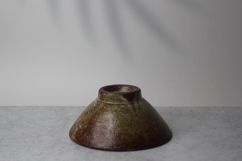 Puede incluir: Un cuenco de cer&aacute;mica hecho a mano con una forma c&oacute;nica invertida inusual. El cuenco presenta un borde marr&oacute;n oscuro y un degradado de tonos terrosos, incluyendo marr&oacute;n y verde oliva. El cuenco probablemente se utiliza para servir o exhibir.