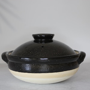 Peut inclure: Un pot japonais en c&eacute;ramique noire, un donabe, avec son couvercle assorti. Le pot a une finition mouchet&eacute;e et une base de couleur cr&egrave;me. Il comporte deux poign&eacute;es et un petit bouton sur le couvercle. Un r&eacute;cipient de cuisson traditionnel.