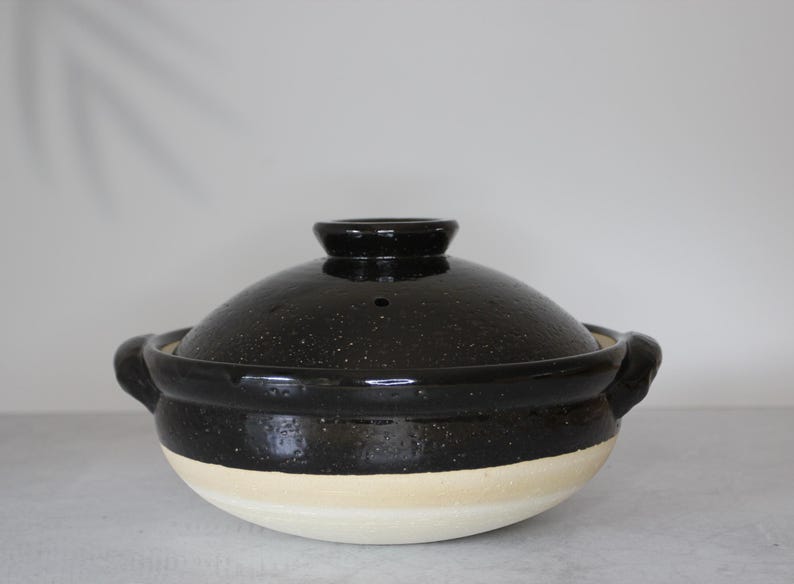 Peut inclure: Un pot japonais en c&eacute;ramique noire, un donabe, avec son couvercle assorti. Le pot a une finition mouchet&eacute;e et une base de couleur cr&egrave;me. Il comporte deux poign&eacute;es et un petit bouton sur le couvercle. Un r&eacute;cipient de cuisson traditionnel.