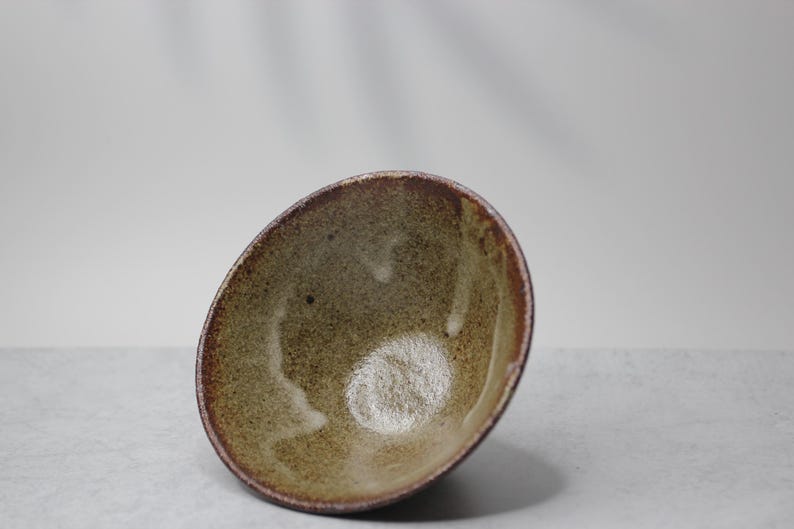 Puede incluir: Un cuenco de cer&aacute;mica hecho a mano con una forma org&aacute;nica. El cuenco presenta un interior moteado de color verde oliva y un borde marr&oacute;n oscuro. El exterior tiene un aspecto texturizado y terroso. Ideal para servir o como pieza decorativa. Medidas en cent&iacute;metros.