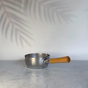 Puede incluir: Un cazo de metal de color plateado que presenta una textura martillada distintiva en su superficie exterior. El recipiente de cocina está equipado con un mango cilíndrico de madera de color marrón claro unido al lateral con un conector metálico.