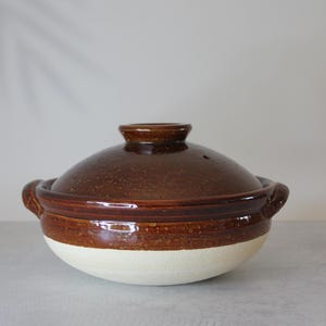 Peut inclure: Une marmite de cuisine en c&eacute;ramique marron avec un couvercle. Le pot a une base blanche et un dessus et un couvercle marron fonc&eacute;. Le couvercle a une petite poign&eacute;e et un trou d'a&eacute;ration. Le pot a deux poign&eacute;es lat&eacute;rales.