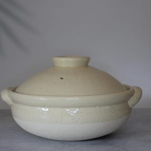 Peut inclure: Pot de cuisson en c&eacute;ramique cr&egrave;me avec couvercle. Le pot a une forme arrondie avec deux poign&eacute;es et un couvercle avec un petit bouton. Le pot est beige clair avec une surface textur&eacute;e, adapt&eacute; &agrave; la cuisine.