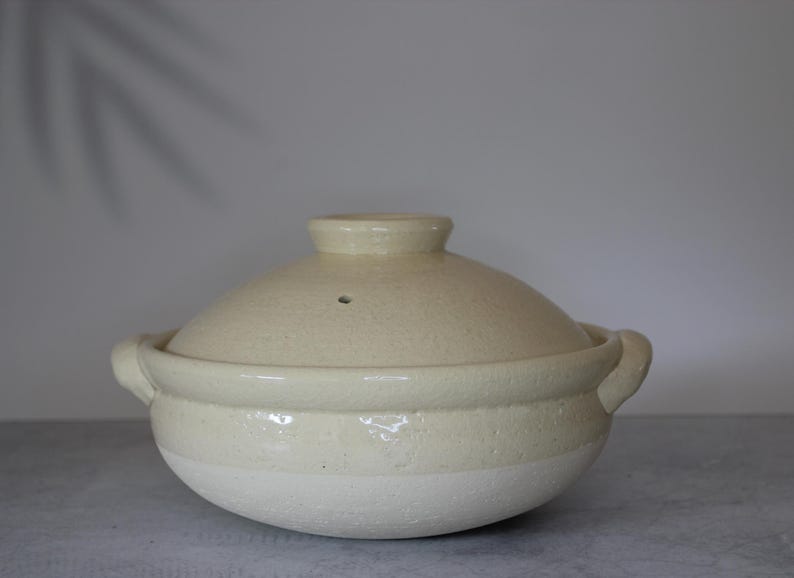 Peut inclure: Pot de cuisson en c&eacute;ramique cr&egrave;me avec couvercle. Le pot a une forme arrondie avec deux poign&eacute;es et un couvercle avec un petit bouton. Le pot est beige clair avec une surface textur&eacute;e, adapt&eacute; &agrave; la cuisine.