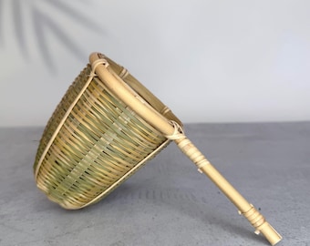 May include: A conical tea strainer crafted from woven bamboo strips, featuring a natural light tan and pale green color palette. The strainer has a sturdy bamboo rim and a long, slender bamboo handle reinforced with thin, tightly wrapped bamboo cord.