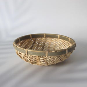 May include: A shallow, round woven basket featuring a natural light tan bamboo or reed construction with a contrasting dark olive green band encircling the upper rim. The basket has an open lattice weave pattern and a sturdy, smooth circular frame.
