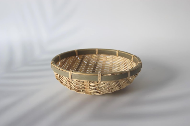 May include: A shallow, round woven basket featuring a natural light tan bamboo or reed construction with a contrasting dark olive green band encircling the upper rim. The basket has an open lattice weave pattern and a sturdy, smooth circular frame.
