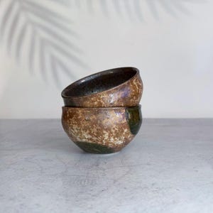 May include: Two stacked ceramic bowls featuring a mottled brown and tan textured glaze with dark green accents. The bowls have a rounded shape with a smooth interior and a rough, earthy exterior finish, showcasing a handcrafted appearance.
