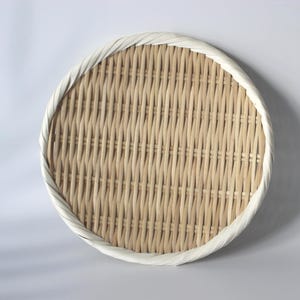May include: A round, flat bamboo basket with a light tan woven base and a thick, white, twisted rim. The basket features a tight, uniform weave pattern throughout the centre, creating a circular tray with a clean, natural aesthetic.
