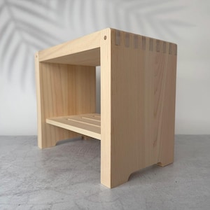 Puede incluir: Un taburete de madera de color claro con una superficie rectangular y un estante inferior de listones. La estructura presenta juntas de dedos visibles en las esquinas, mostrando un acabado de veta natural y un diseño robusto.