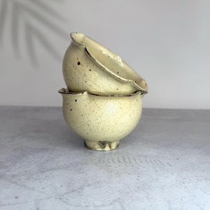 May include: Two stacked ceramic bowls with a speckled cream-coloured glaze and a dark brown rim. The bowls feature a rounded, organic shape with a scalloped edge design and a small footed base, showcasing a textured finish with visible dark brown spots.