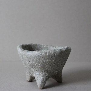 Puede incluir: Un pequeño macetero de cerámica gris con una superficie rugosa y texturizada. El macetero tiene tres patas y una parte superior redondeada en forma de cuenco.