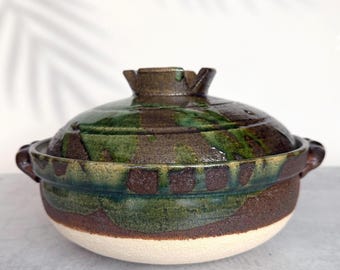 May include: A round ceramic lidded pot featuring a textured, unglazed beige base and a dark brown upper section with a glossy, deep green drip glaze. The lid has a matching dark brown and green finish with a central knob and two side handles.